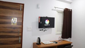 A room with a wooden door, a wall-mounted TV, a desk with a kettle, a remote, and a bottle. - Anandam Grand (Greater Noida)
