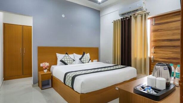 A hotel room with a wooden bed, a nightstand with a vase of flowers, a coffee maker, and a wardrobe. - Anandam Grand (Greater Noida)