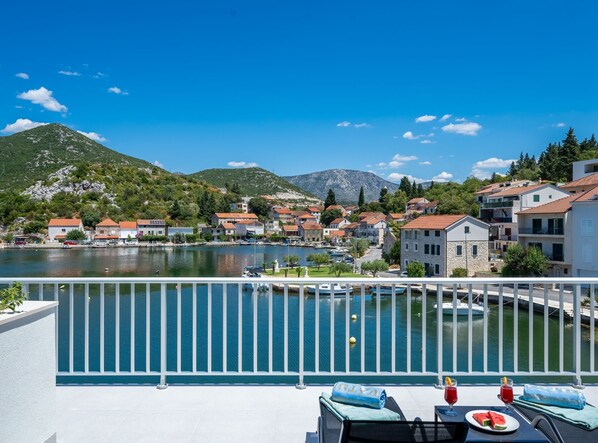 A balcony with a view of a lake, boats, and a hillside village. - Villa Senka (Ploce)
