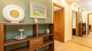 A well-lit hallway with a wooden console table displaying a decorative plate, a framed painting, and a small vase. A wooden door and a mirror are also visible.