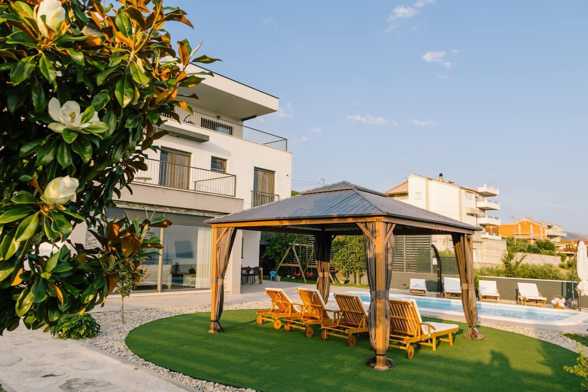 A modern outdoor pool area with lounge chairs, a covered seating area, and a building with balconies.