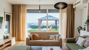 Un soggiorno moderno con un divano beige, una poltrona verde e un tavolino da caffè. Un'ampia finestra si affaccia su un giardino e un gazebo.