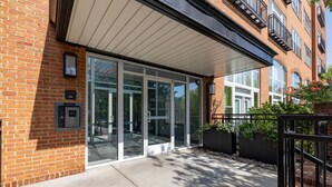 A modern building entrance with glass doors, a brick facade, and a small outdoor seating area with plants. - Downtown Raleigh, Pet Friendly, Close to everything. (Raleigh)