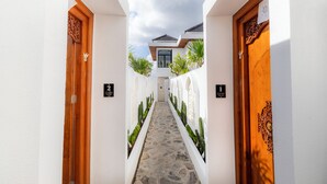 A narrow hallway with white walls, wooden doors, and a stone floor leading to a building with a sloped roof. - Kamaimo Villa (Munggu)