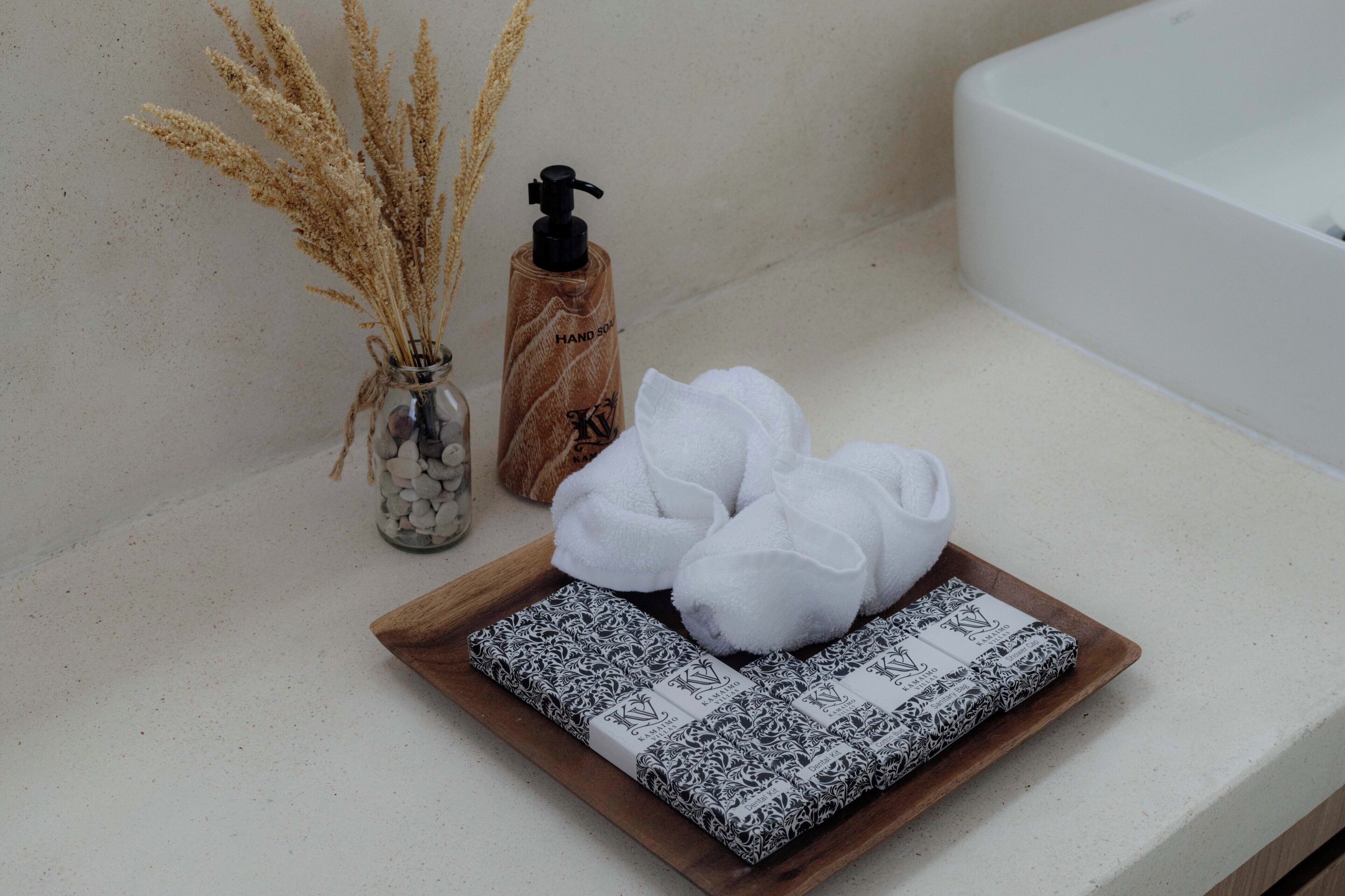A bathroom counter with a glass vase of dried flowers, a bottle of hand soap, and a wooden tray with rolled towels and a patterned box.
