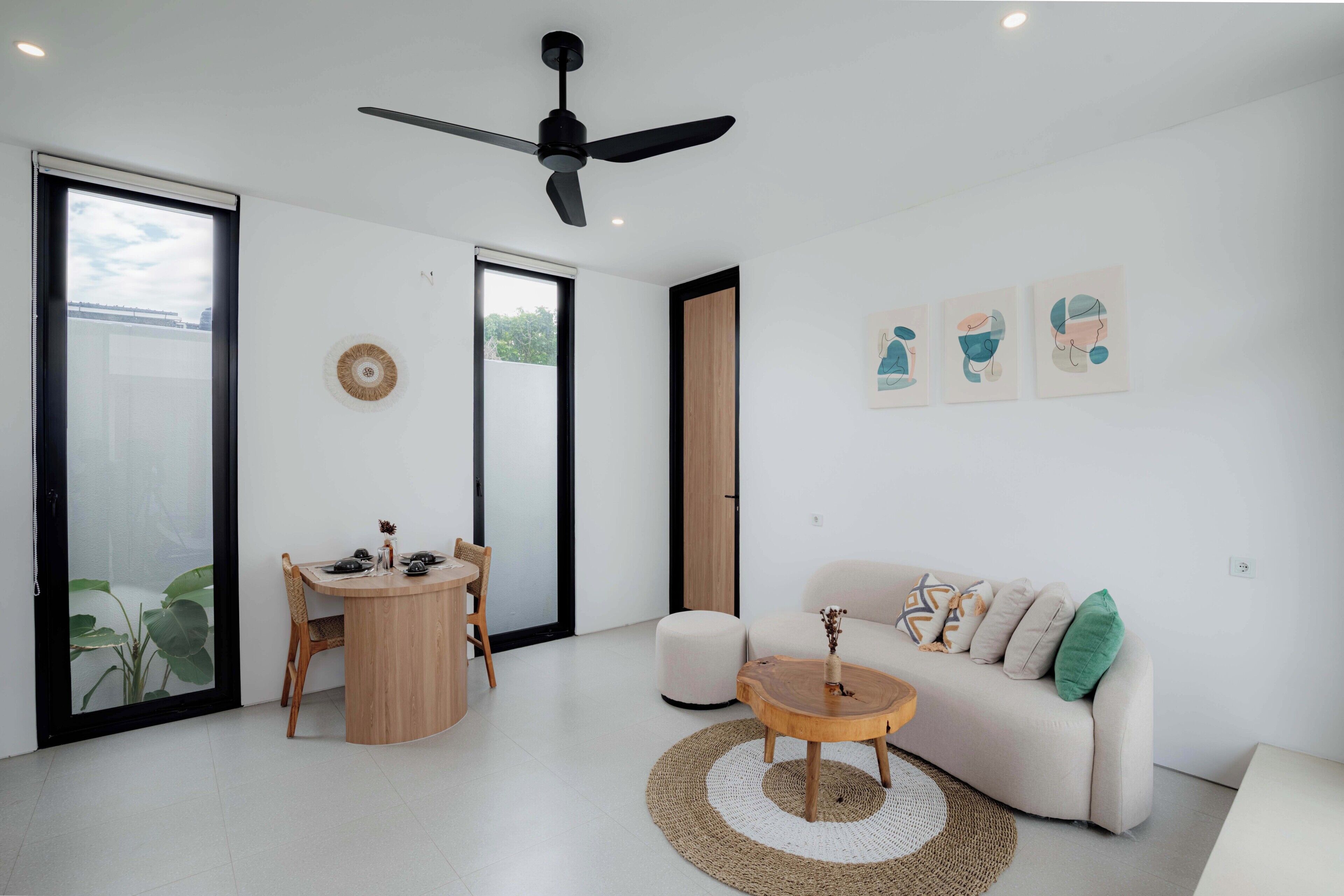 Un soggiorno moderno con un tavolo rotondo in legno, un divano bianco con cuscini, una ventola a soffitto e un'ampia finestra con vista.