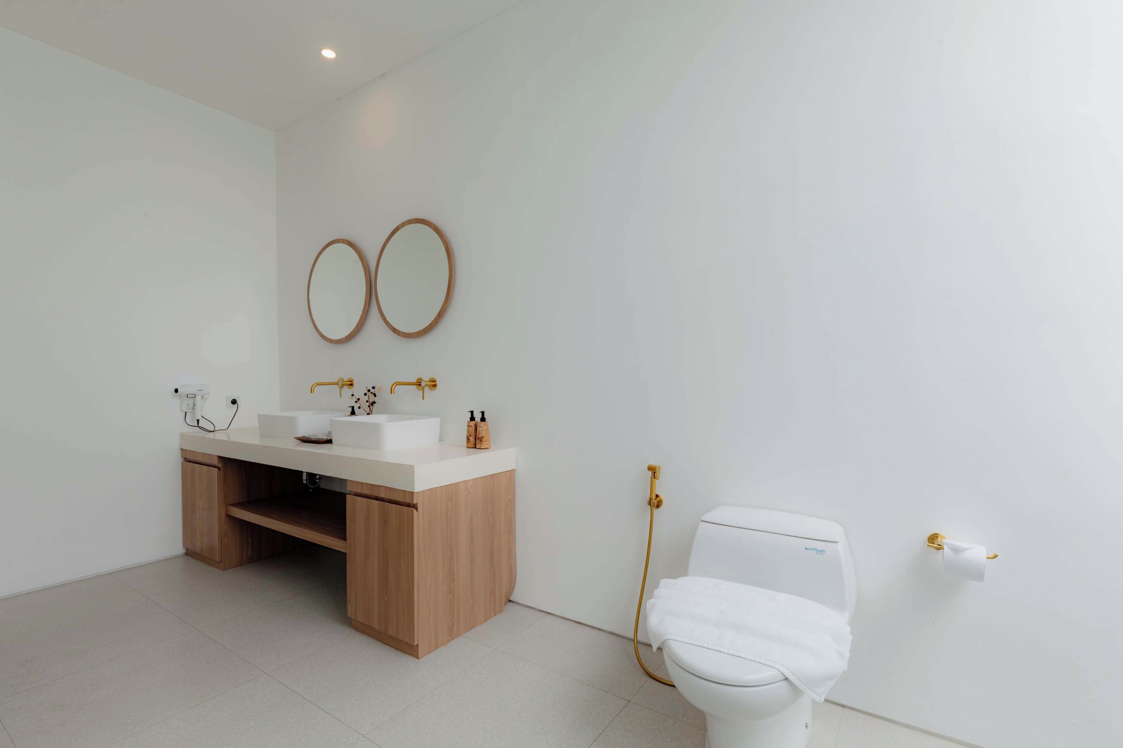 Un baño moderno con un tocador de madera, dos espejos redondos, un inodoro blanco y una ducha con detalles dorados.