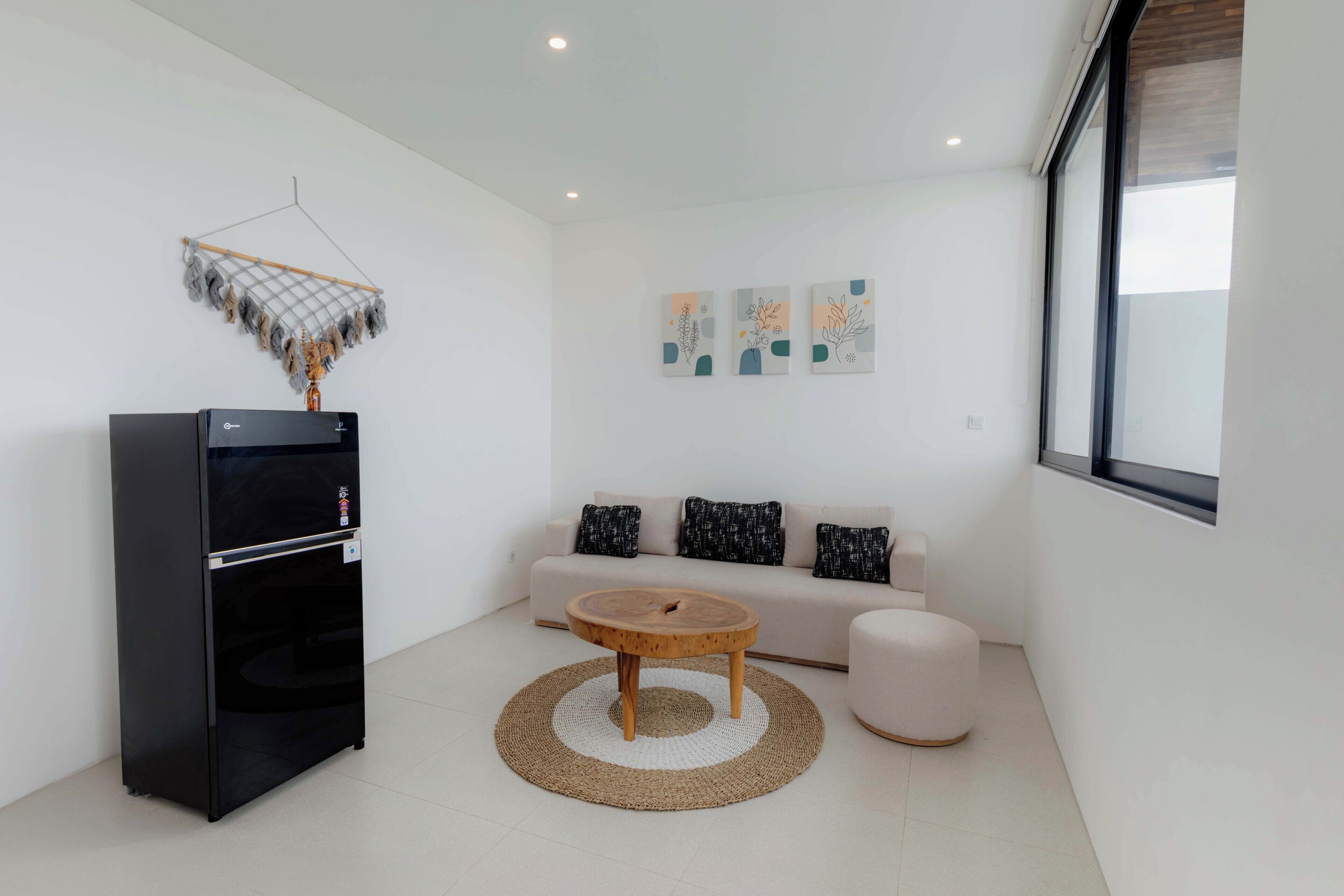 A modern living room with a black refrigerator, a round wooden coffee table, a beige sofa with patterned cushions, and abstract wall art.