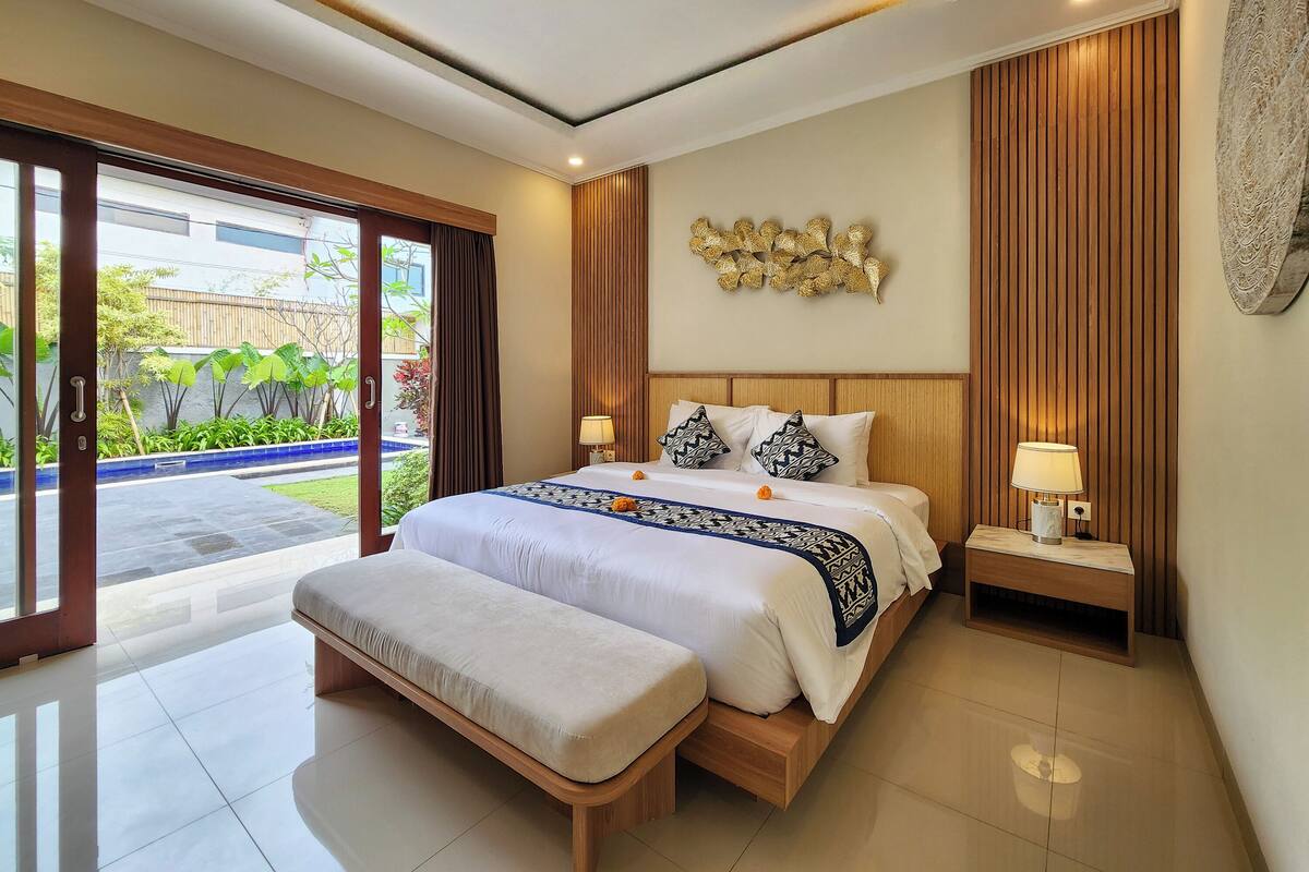 A modern bedroom with a large bed, a bench, and a view of a garden through a sliding door.
