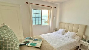 A bedroom with a bed, a reading area with a book, a window with a view of the sea, and a nightstand with a lamp. - Casa Ale, endless sea view ! (Las Aguas)