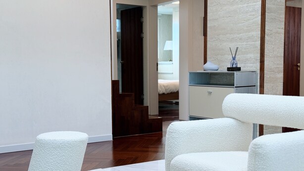 Una habitación de hotel moderna con una cama grande, un escritorio pequeño y un área de descanso con sillas blancas acolchadas.