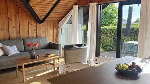 A cozy room with a wooden ceiling, a grey sofa, a wooden table, and a view of greenery outside. - Cozy summer house with new kitchen and bathroom (Karrebæksminde)