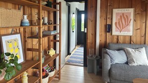 A wooden interior with a bookshelf, a framed picture, a grey sofa, and a door leading outside. - Cozy summer house with new kitchen and bathroom (Karrebæksminde)