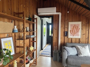 Ein Holzinterieur mit Bücherregal, gerahmtem Bild, grauem Sofa und einer zur Außenwelt führenden Tür.