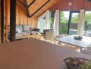 A cozy room with a wooden ceiling, a sofa, a dining table, and a view of greenery outside.