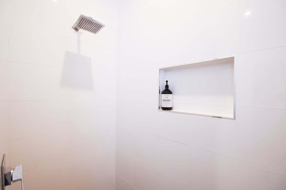 Un baño moderno con azulejos blancos, un cabezal de ducha y una botella de vino sobre una estantería.