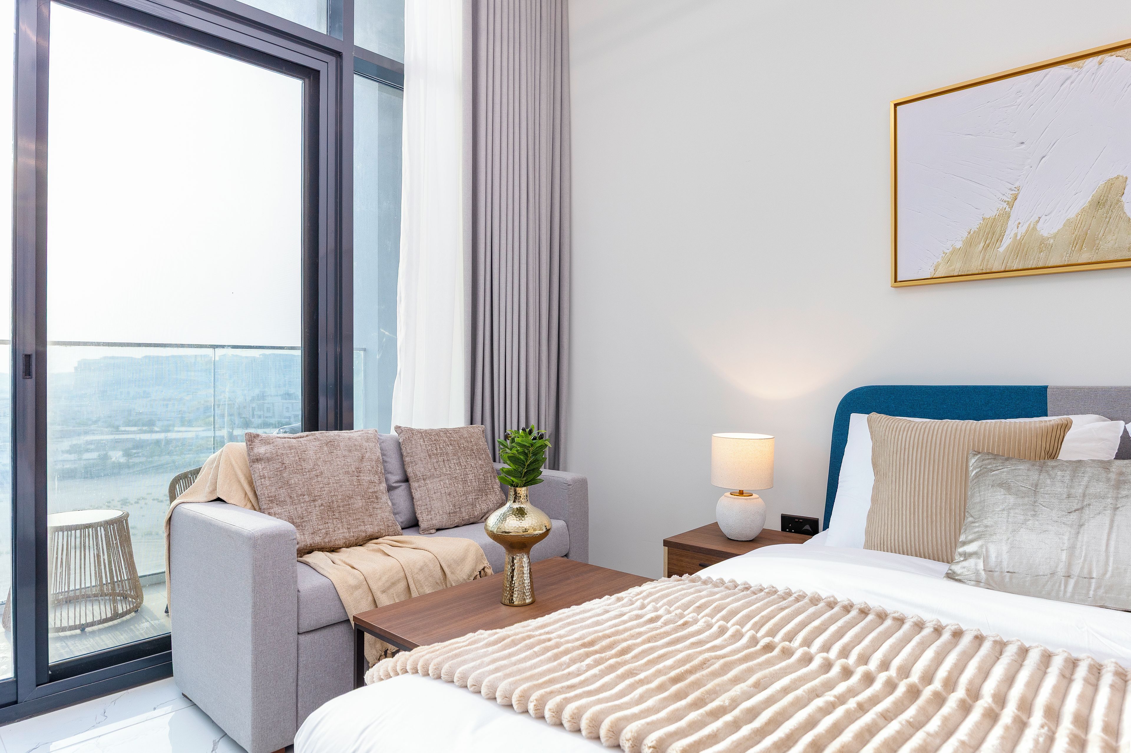 A modern hotel room with a large window, a sofa, a bed, and a nightstand with a lamp.
