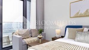 A modern hotel room with a large window, a grey sofa, a wooden side table, a white bed, and a blue headboard. - Bloomfields Comfort Stu The Gate  (Abu Dhabi)