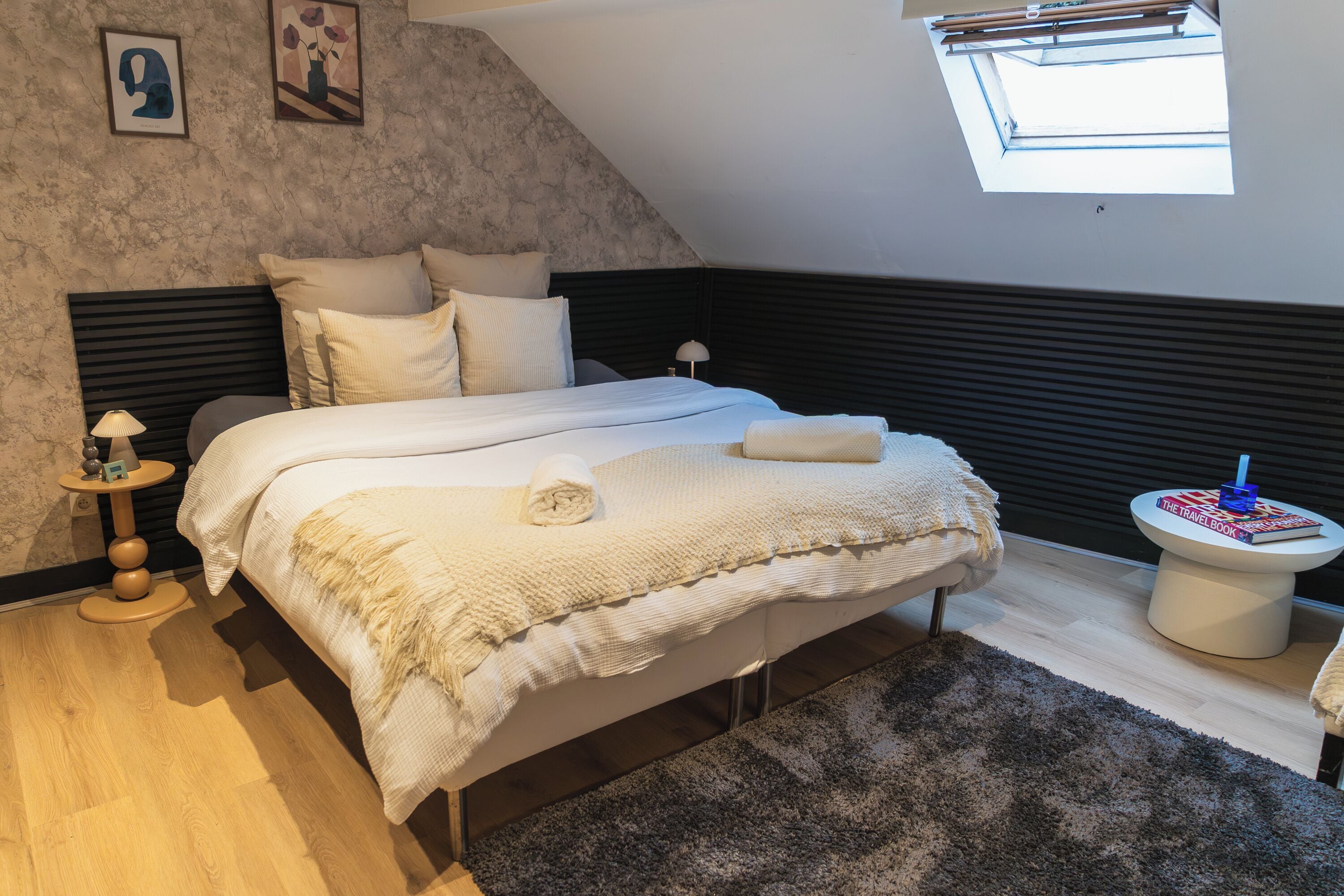 A bedroom with a sloped ceiling, a bed with white bedding, a small bedside table with a lamp, and a window providing natural light.