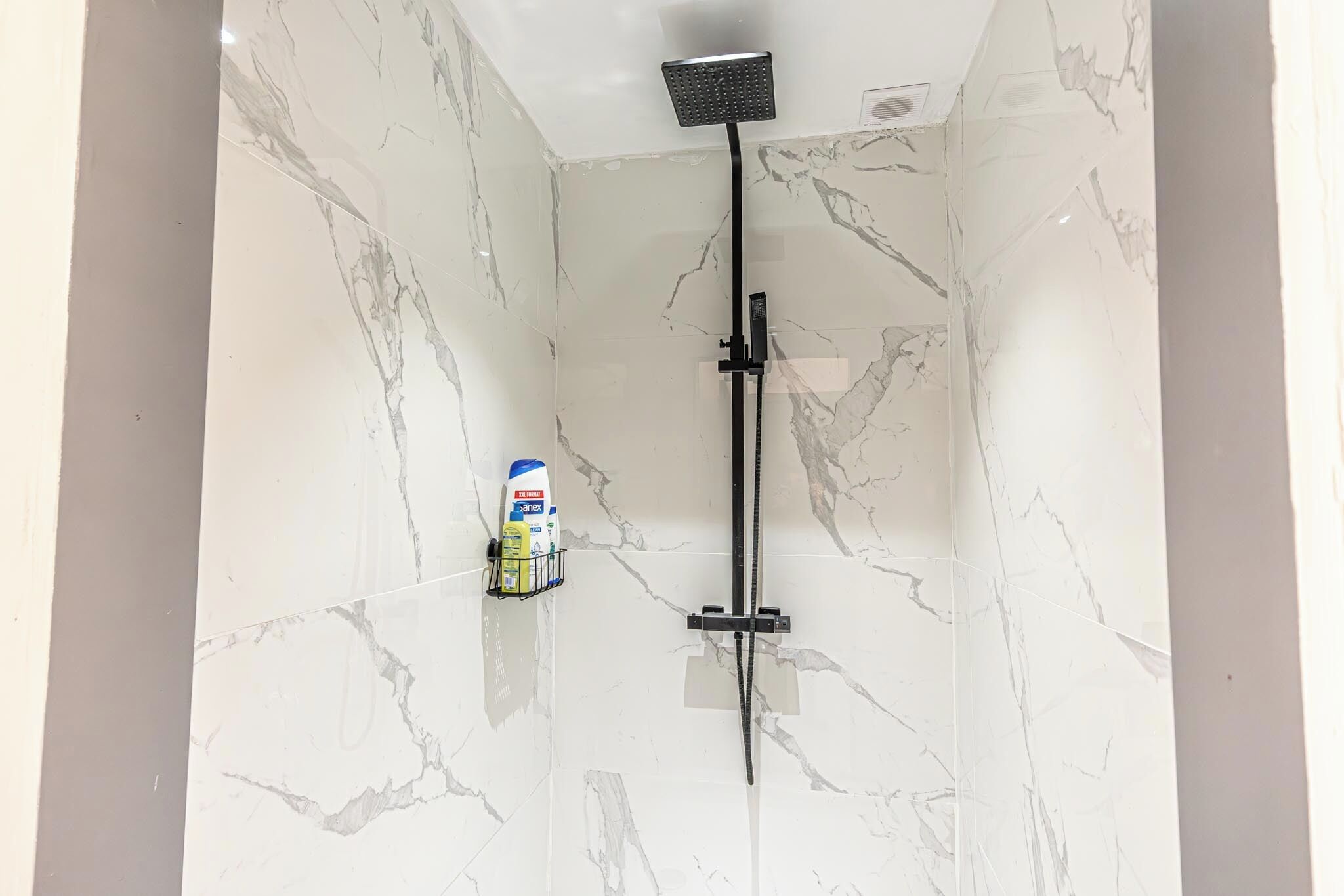 Une salle de bain avec une douche, des murs en marbre et une bouteille de shampoing posée sur une étagère.