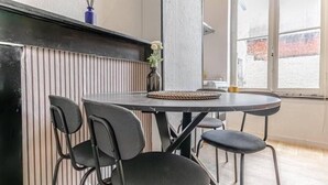 Un comedor con una mesa redonda y cuatro sillas, un florero con flores, y vistas a un edificio de ladrillo a través de una ventana.