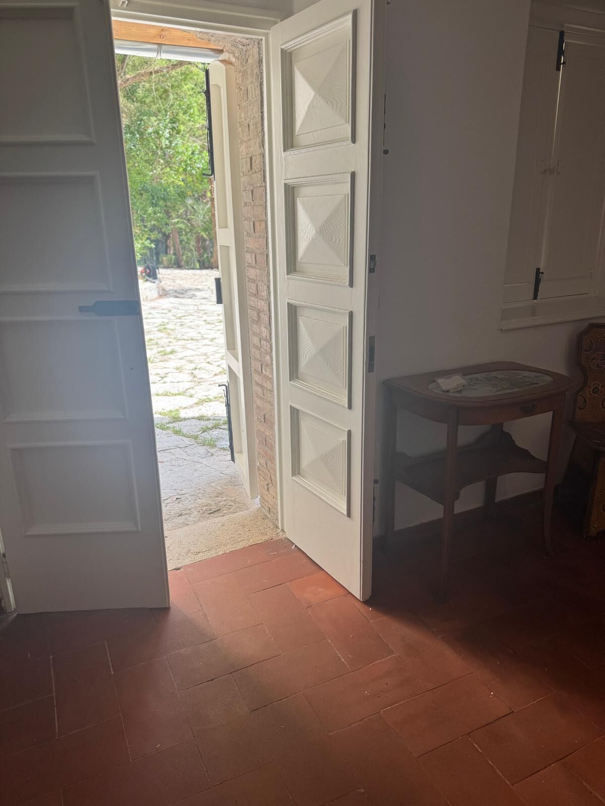 Una habitación con una puerta blanca, una pequeña mesa redonda y vistas a una pared de ladrillo y vegetación exterior.