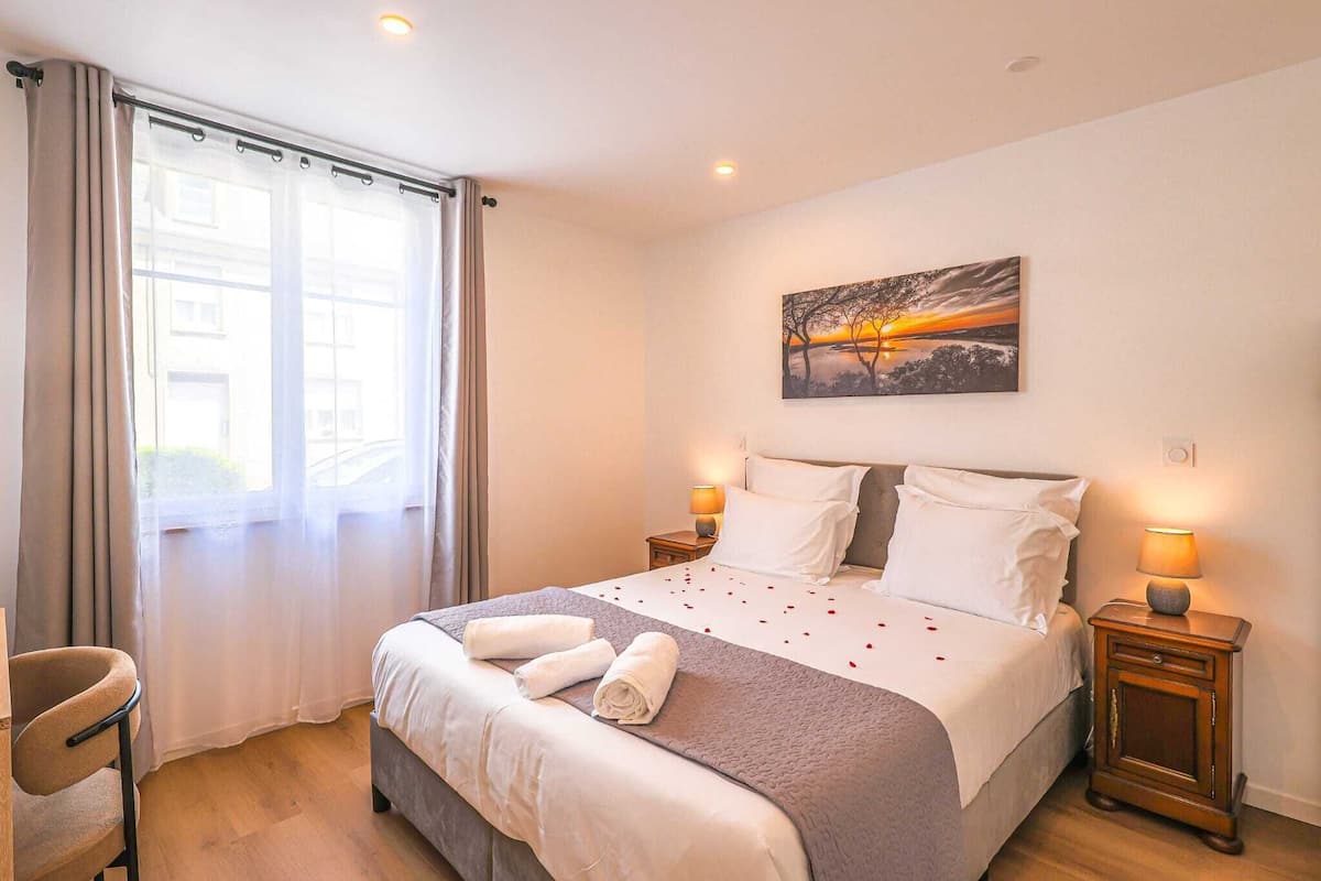 Un lit bien fait, avec du linge de lit blanc et une couverture grise, deux lampes de chevet, un tableau représentant un coucher de soleil et une fenêtre avec des rideaux.