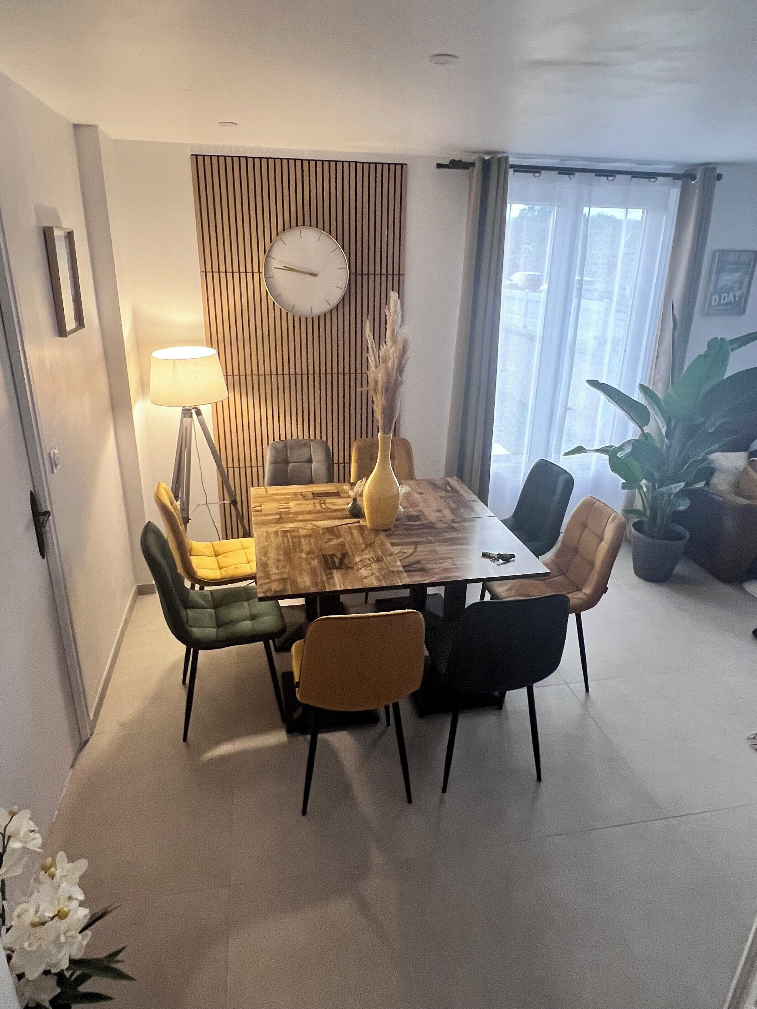 Un coin repas avec une table en bois, six chaises, une horloge au mur et une plante en pot.