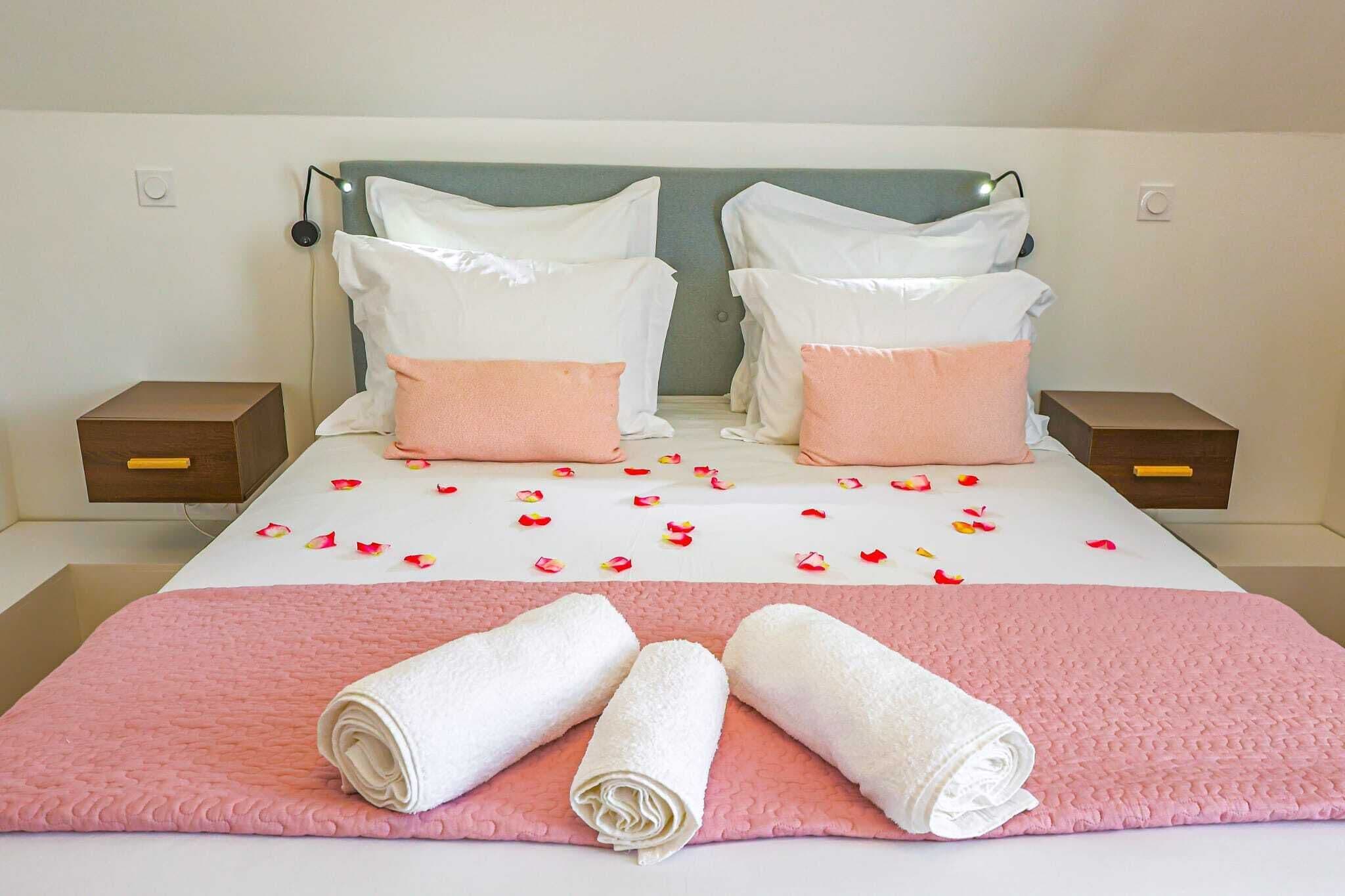Un lit bien fait, avec une literie rose et blanche, deux oreillers et des serviettes blanches roulées posées sur une couverture rose.