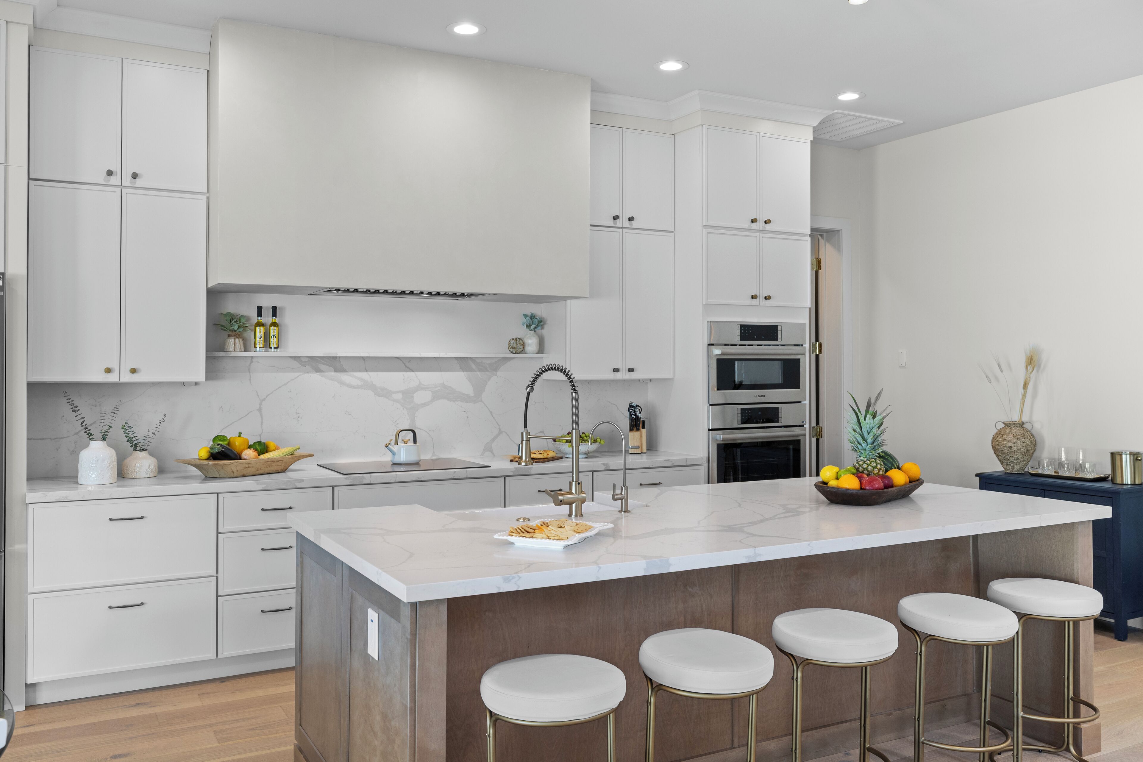 A modern kitchen with white cabinetry, a marble island with stools, and a variety of appliances including a stove, oven, and microwave.