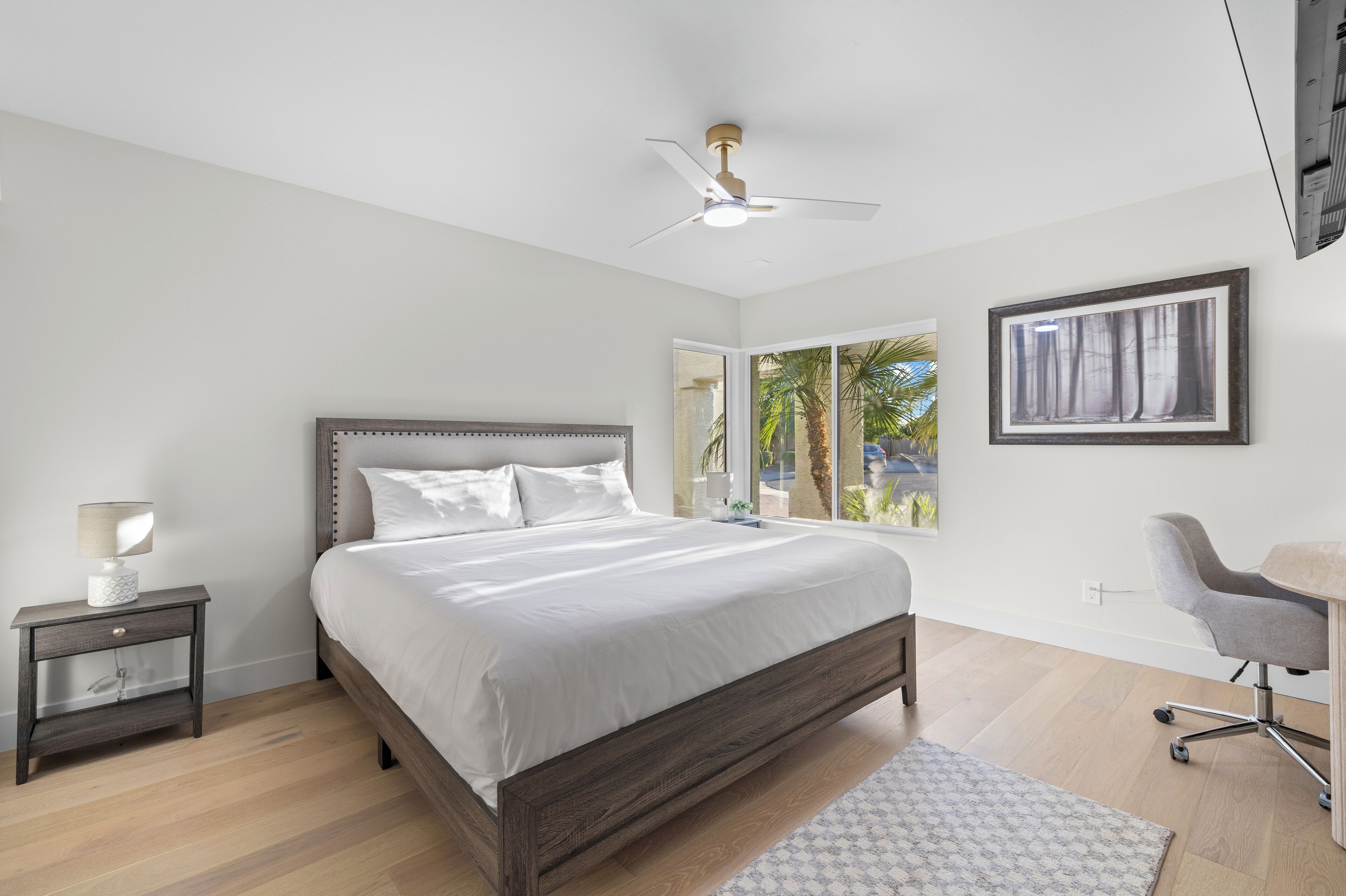 A bedroom with a bed, a nightstand, a desk, a chair, a ceiling fan, and a window with a view of palm trees.