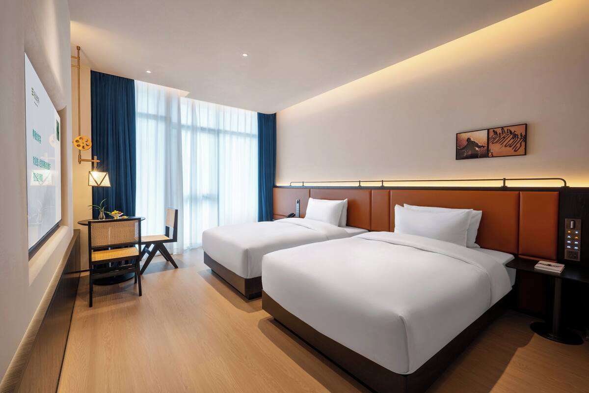 Une chambre d’hôtel avec deux lits, un bureau, une chaise, une lampe et un panneau de commande fixé au mur.