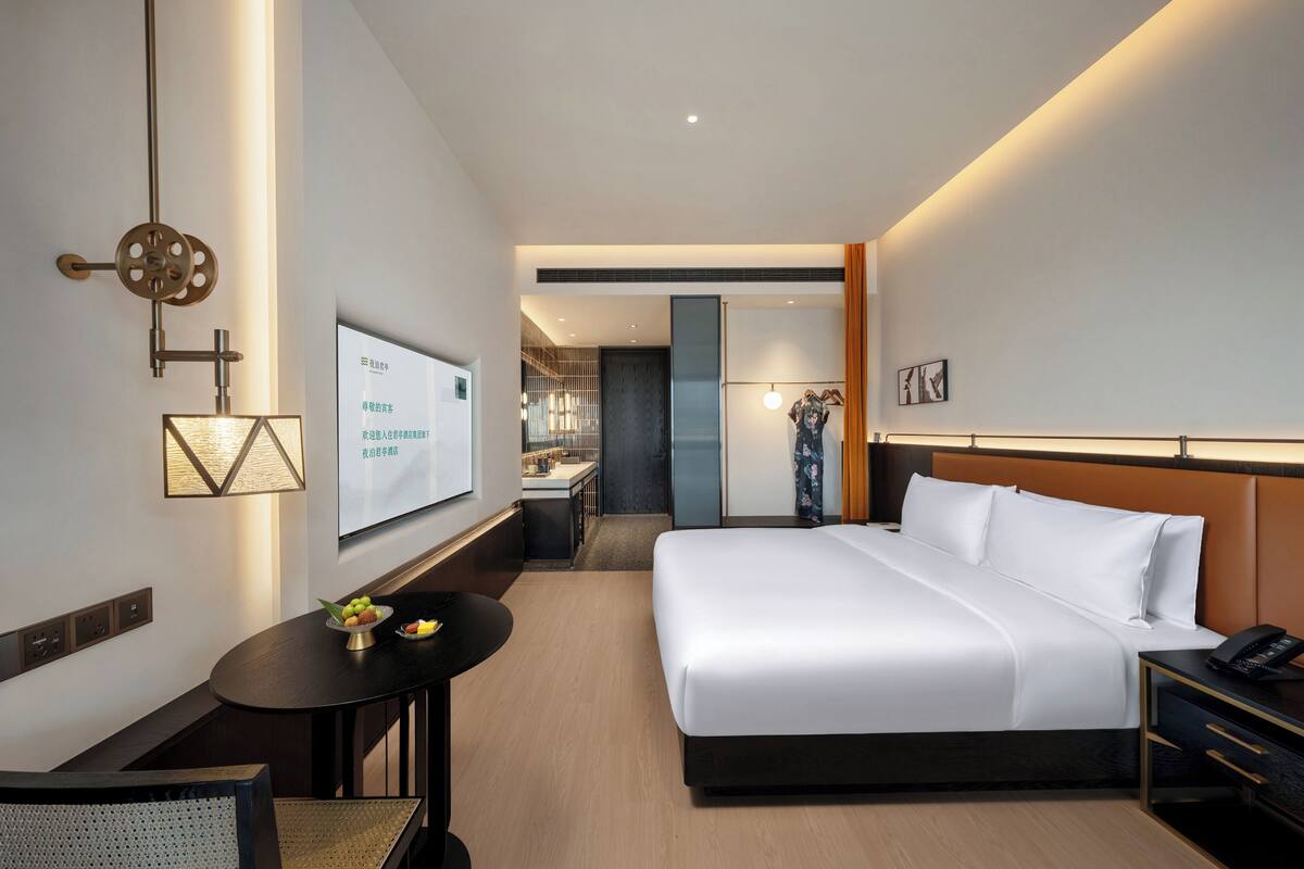 Une chambre d’hôtel moderne avec un grand lit, un bureau avec une chaise, une petite table avec des fruits et un téléviseur fixé au mur.