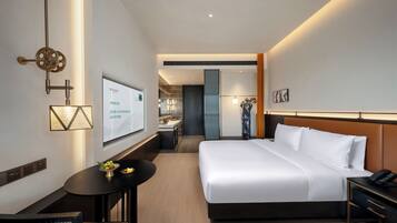 A modern hotel room with a large bed, a desk with a chair, a small table with fruit, and a TV on the wall.