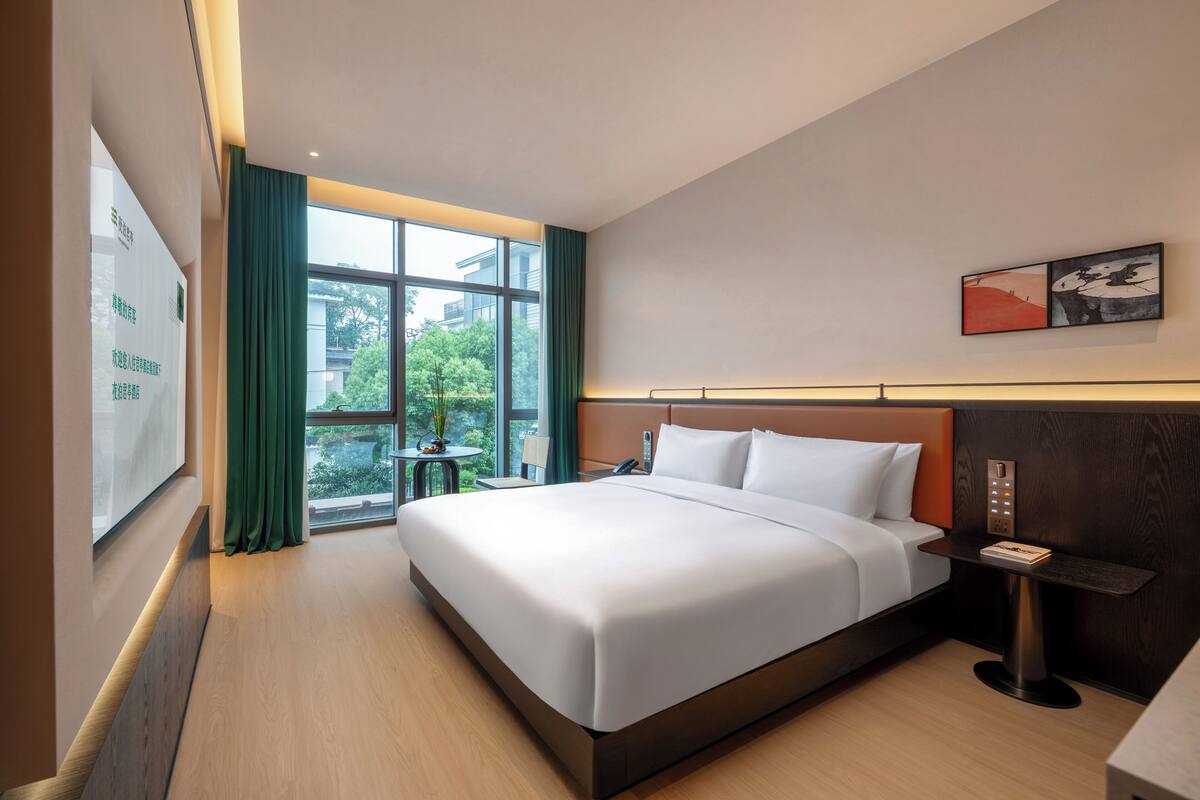 Une chambre d’hôtel avec un grand lit, une table de chevet avec une lampe, une fenêtre avec des rideaux et une vue sur la verdure.