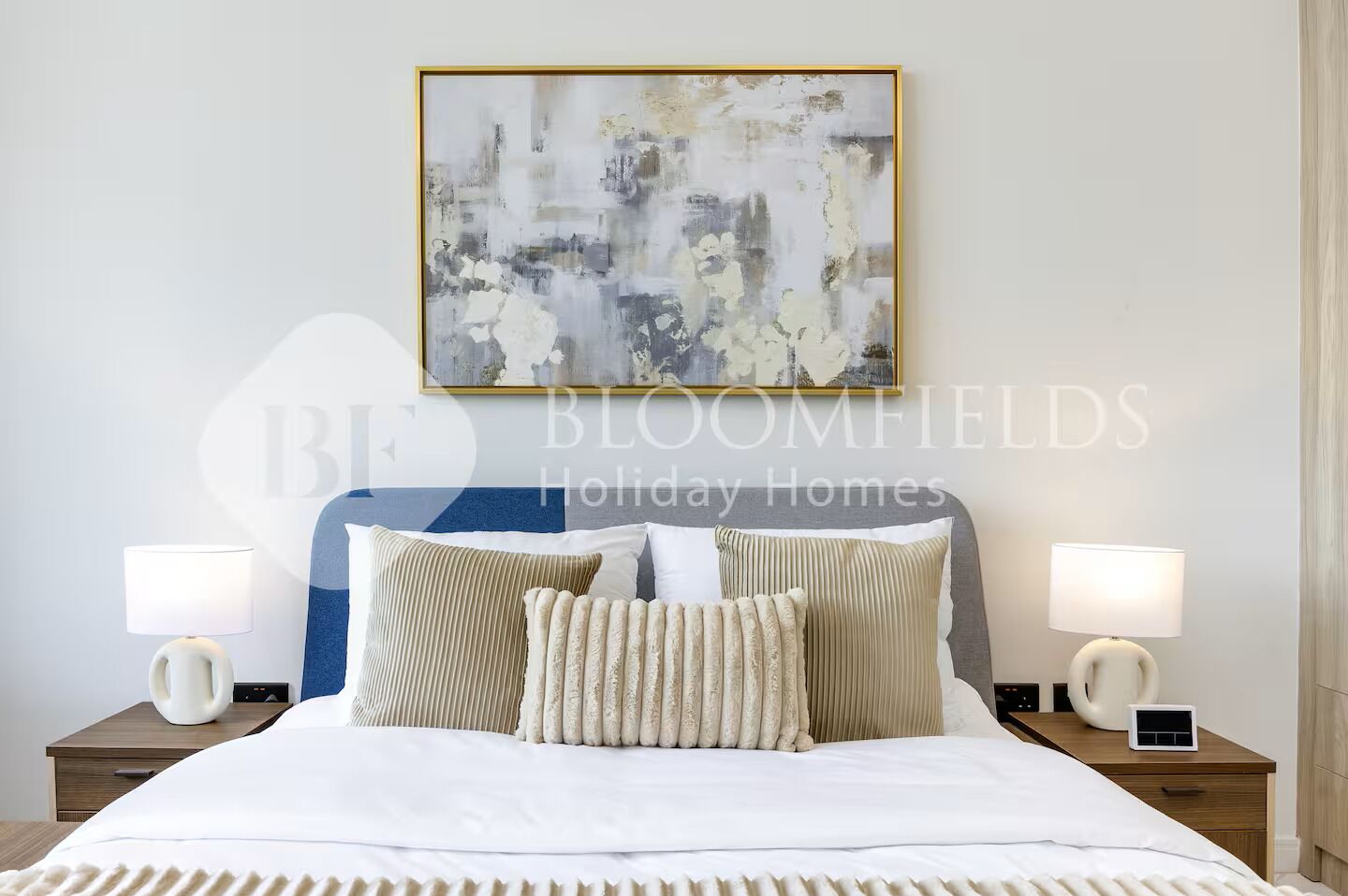A neatly made bed with a blue headboard, two bedside lamps, and a framed abstract painting above it.
