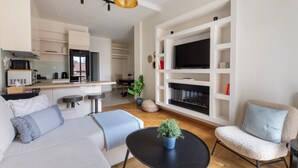 A modern living room with a sofa, armchair, and coffee table, a kitchen area with appliances, and a TV mounted on a white wall. - Attica - Captivating 2-Bedroom Apt & Balcony in Athens (Athens)