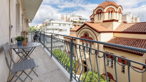 A balcony with a table and chairs, a potted plant, and a view of buildings with a church. - Attica - Captivating 2-Bedroom Apt & Balcony in Athens (Athens)