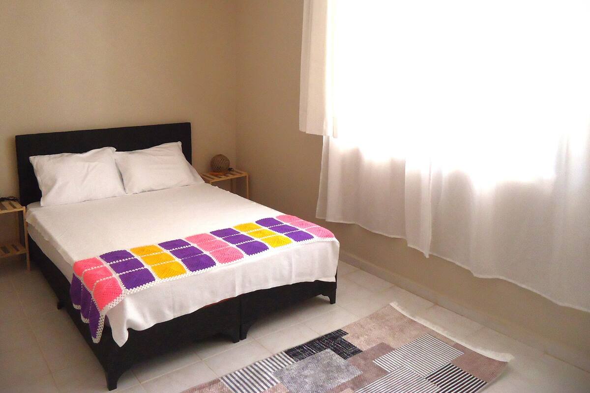 A neatly made bed with a colorful blanket, a bedside table with a lamp, and a window with curtains.