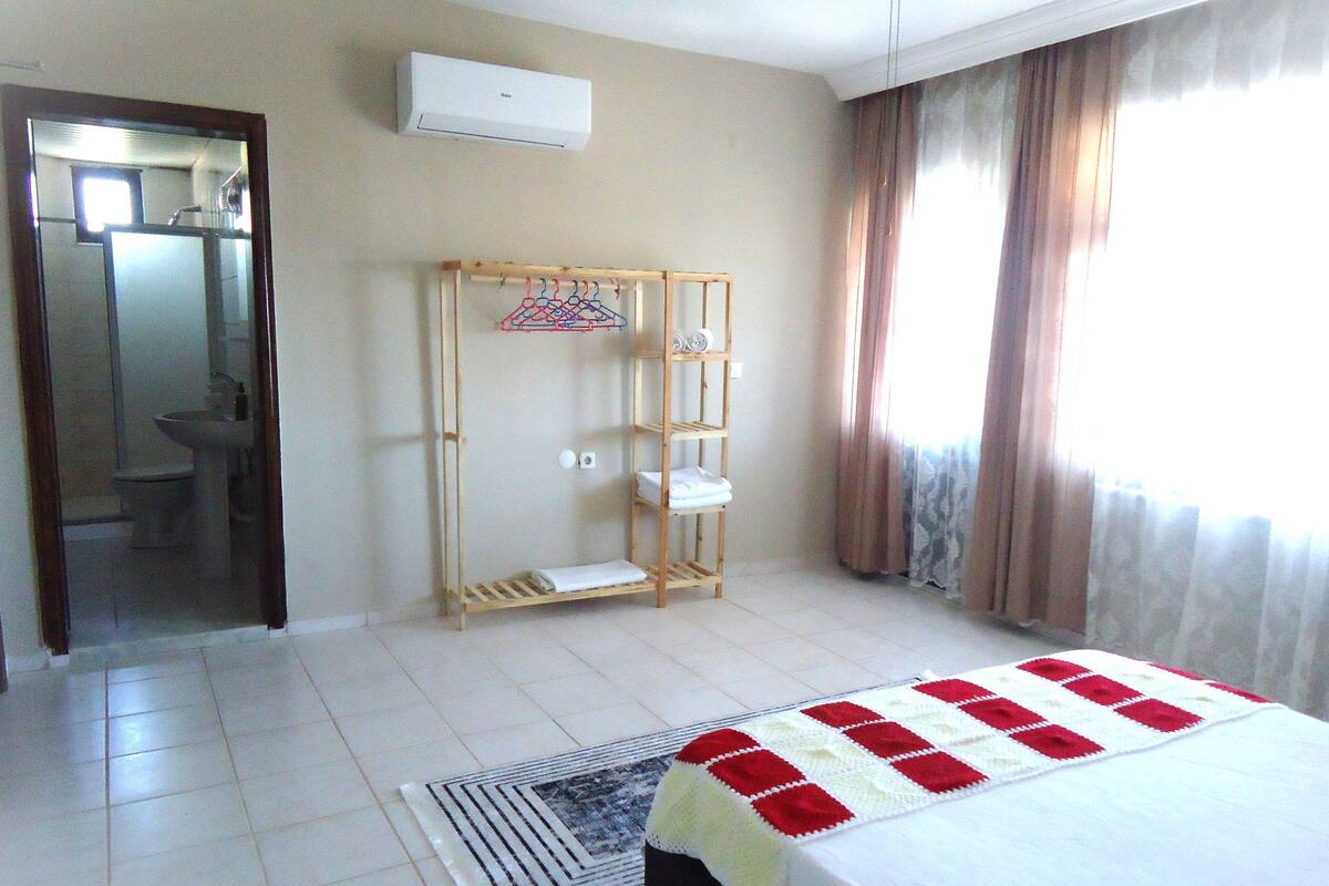 A hotel room with a bed, a wooden shelf unit, a bathroom visible through an open door, and a window with curtains.