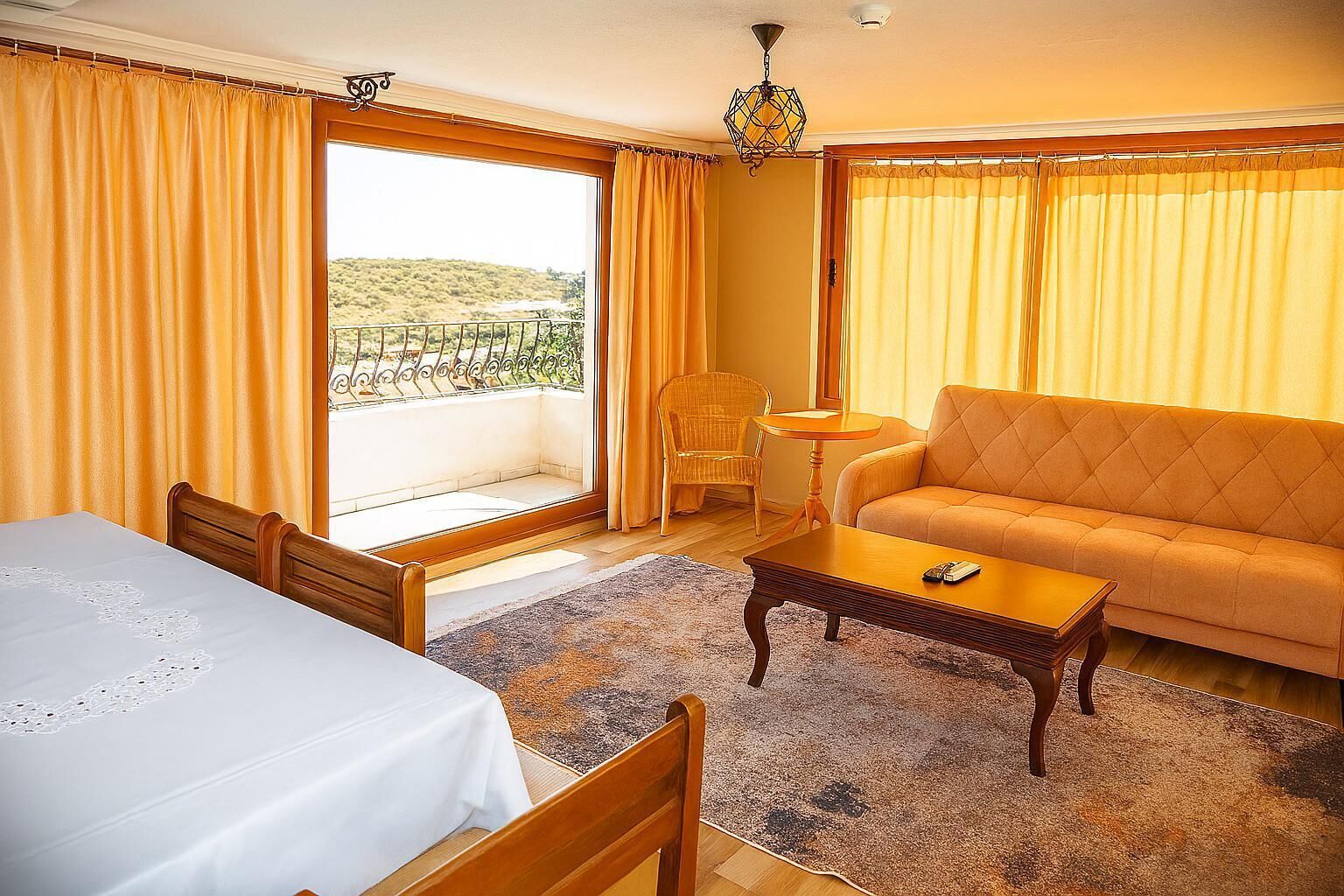A room with a bed, a sofa, a small table, and a chair. There is a window with curtains, a view of the outside, and a chandelier.