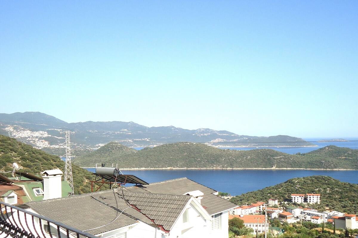 A coastal town with buildings, a solar panel on a roof, and a clear view of the sea and mountains in the background.