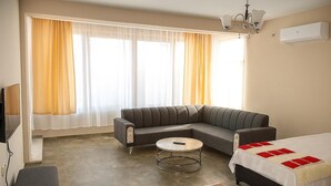 Una sala moderna con un sofá modular, una mesa redonda de centro, una cama con colcha roja y blanca, y un televisor a la izquierda.