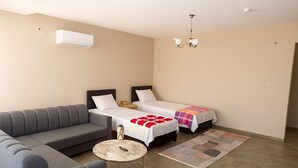Habitación de hotel con un sofá gris, dos camas individuales con ropa blanca, una mesita redonda pequeña, un candelabro y un equipo de aire acondicionado.