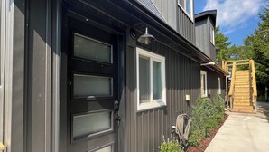 Une maison moderne à deux étages, avec une façade sombre, une porte d’entrée dotée d’un panneau de verre, une petite fenêtre et une terrasse en bois.