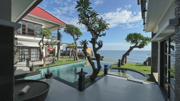 Zona de piscina moderna con vista al mar, rodeada de plantas tropicales y un edificio con tejado rojo.
