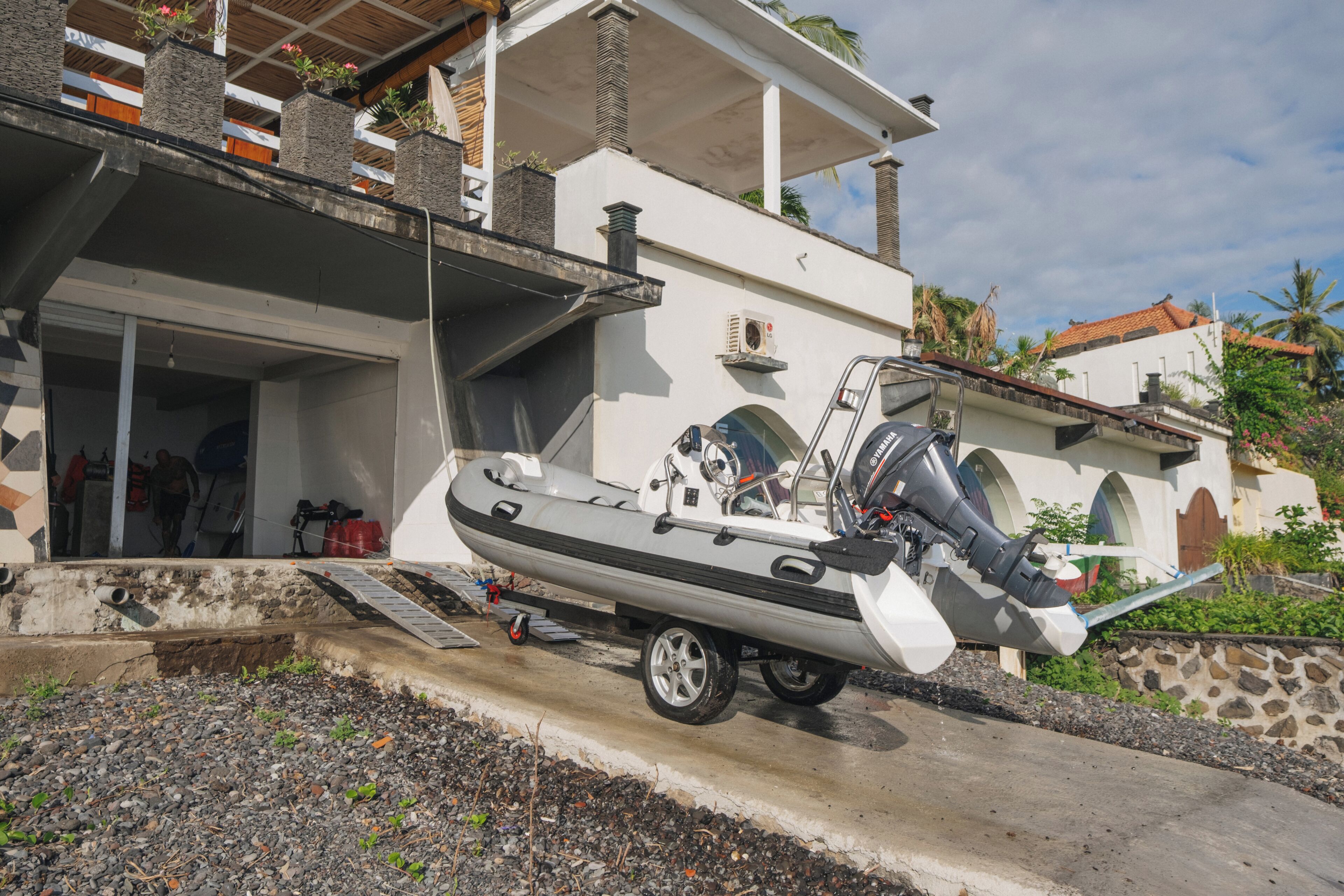 Un bote sobre un remolque estacionado frente a una casa con balcón y un muro de piedra.