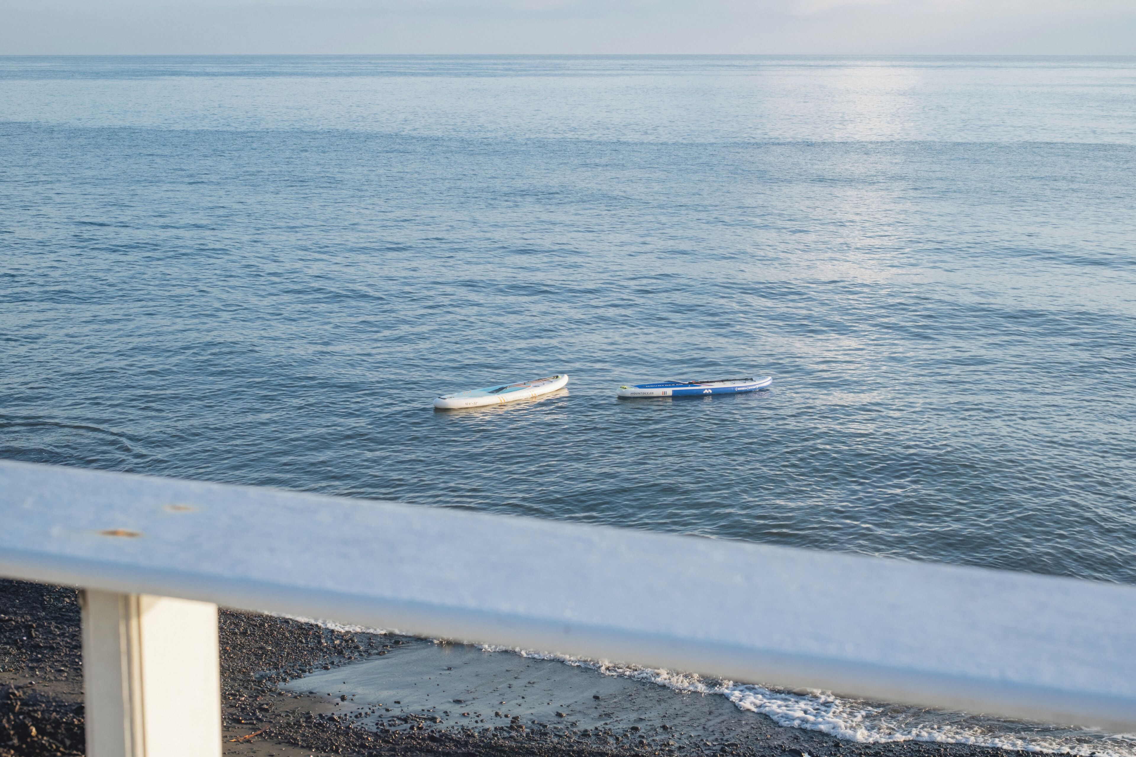 Una playa con dos barcos flotando en agua tranquila, una baranda al frente y una vista despejada del océano.