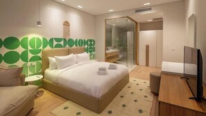 Habitación de hotel moderna con una cama grande, un escritorio, un televisor y un baño visible a través de una puerta de vidrio.