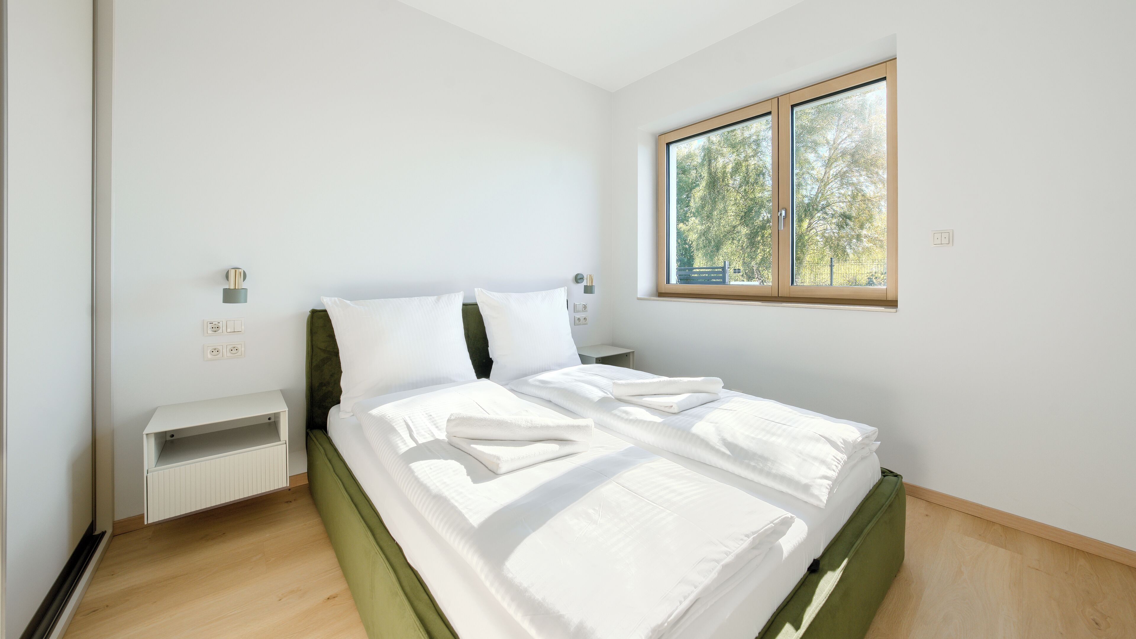 Ein Schlafzimmer mit Bett, Nachttisch, Fenster und Blick auf Bäume.
